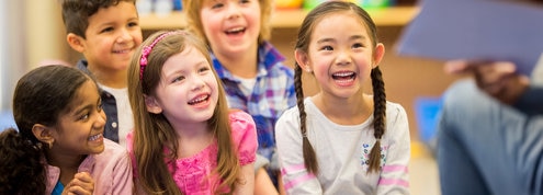 Children smiling and laughing while an adult reads a book to them.