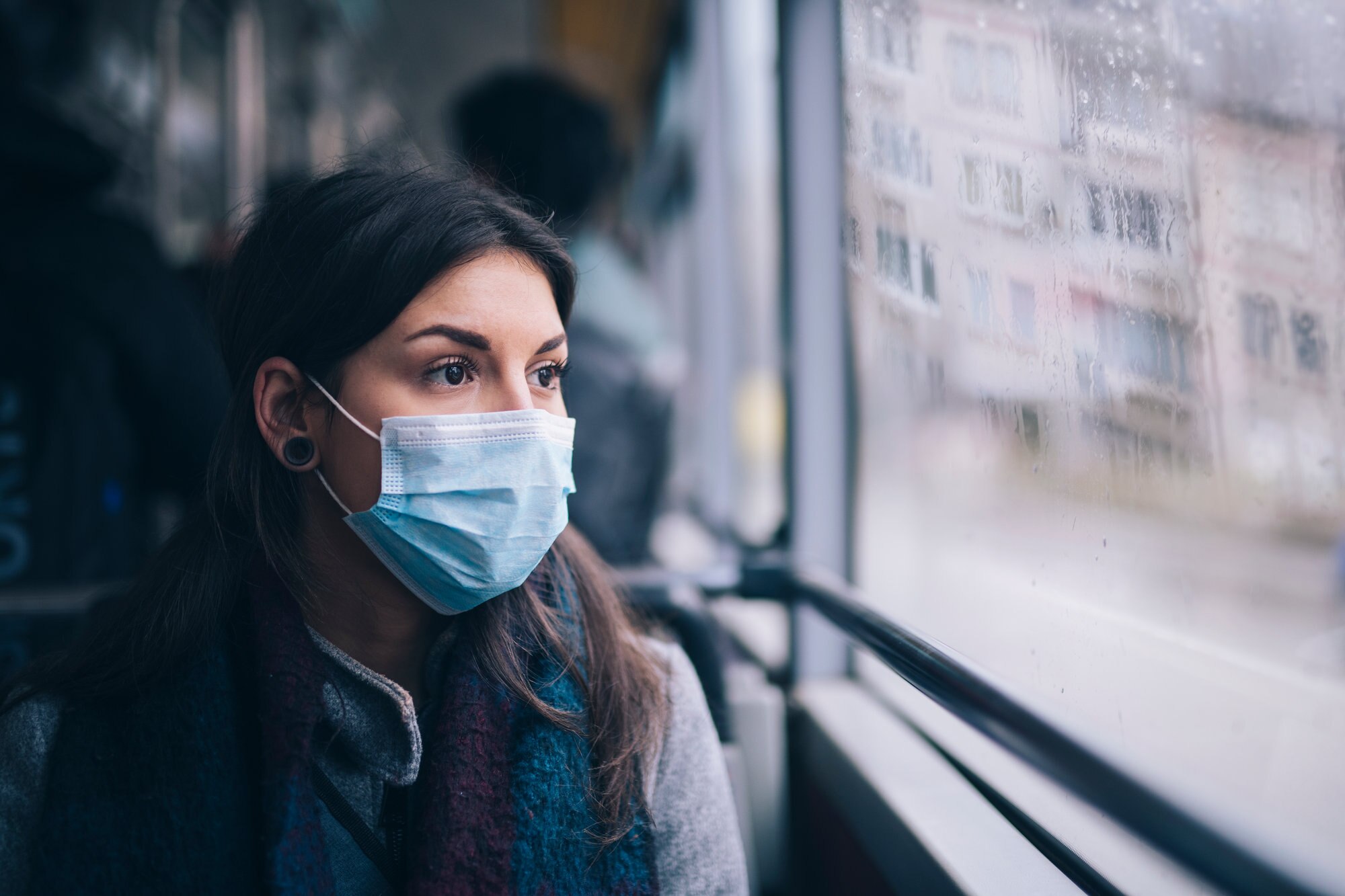A woman sitting on a bus, looking out the window and wearing a protective mask.
